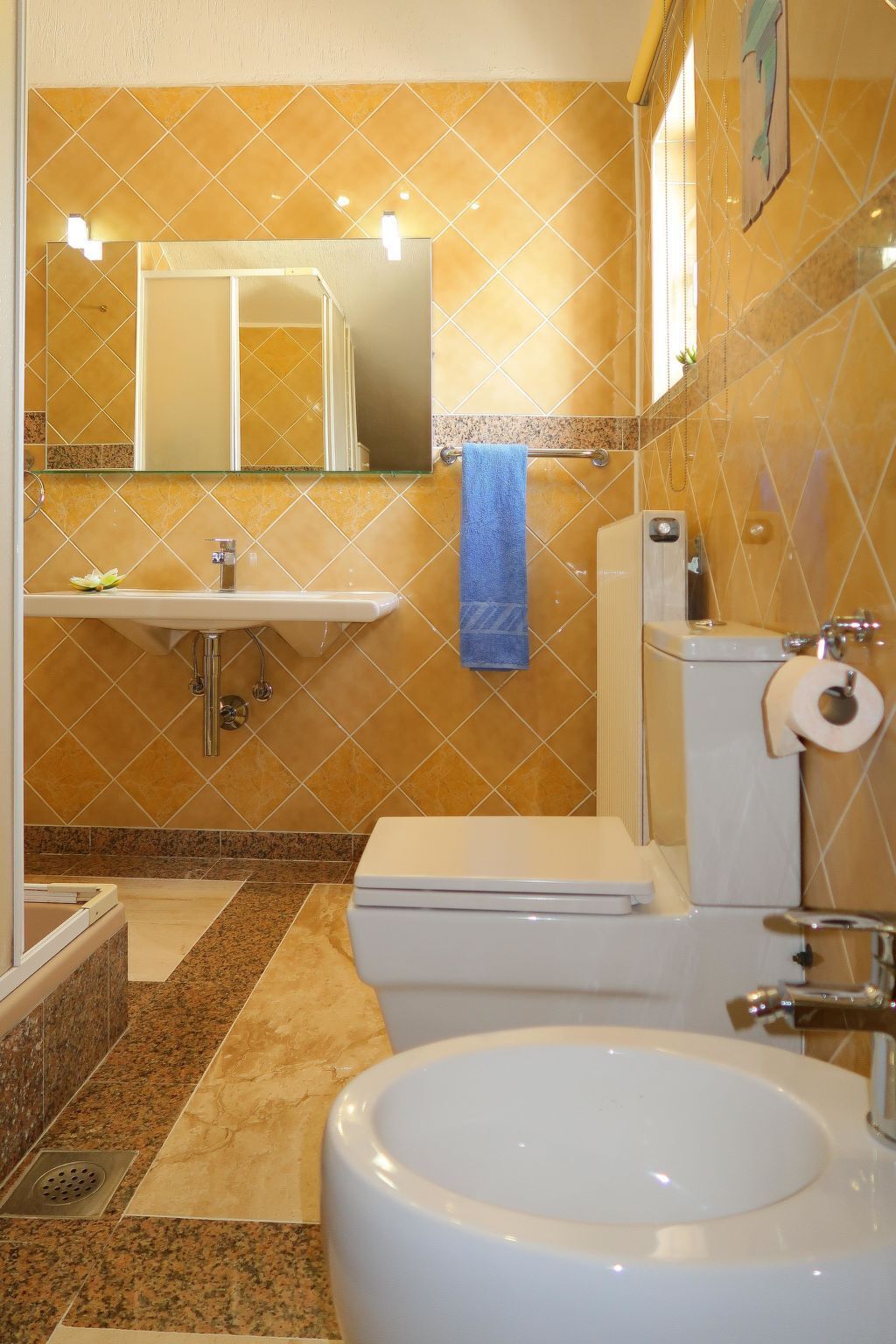 Ein Badezimmer mit Waschbecken, Toilette und Bidet, mit einer gelb gefliesten Wand und einem blauen Handtuch.