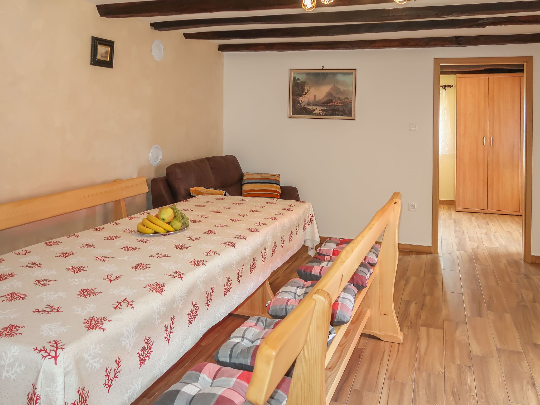 A cozy room with a wooden bench, a dining table with a fruit bowl, a brown sofa, and a painting on the wall.