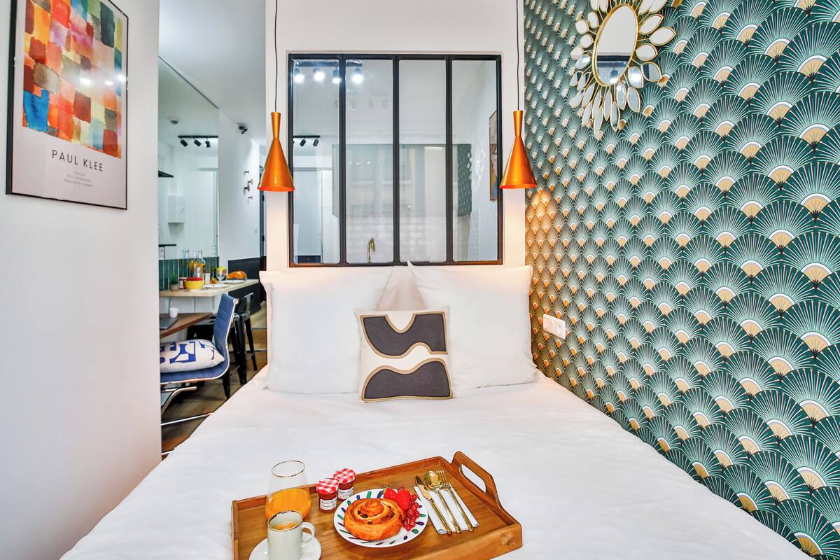 Habitación de hotel ordenada con cama, bandeja de desayuno, ventana y un papel tapiz estampado.