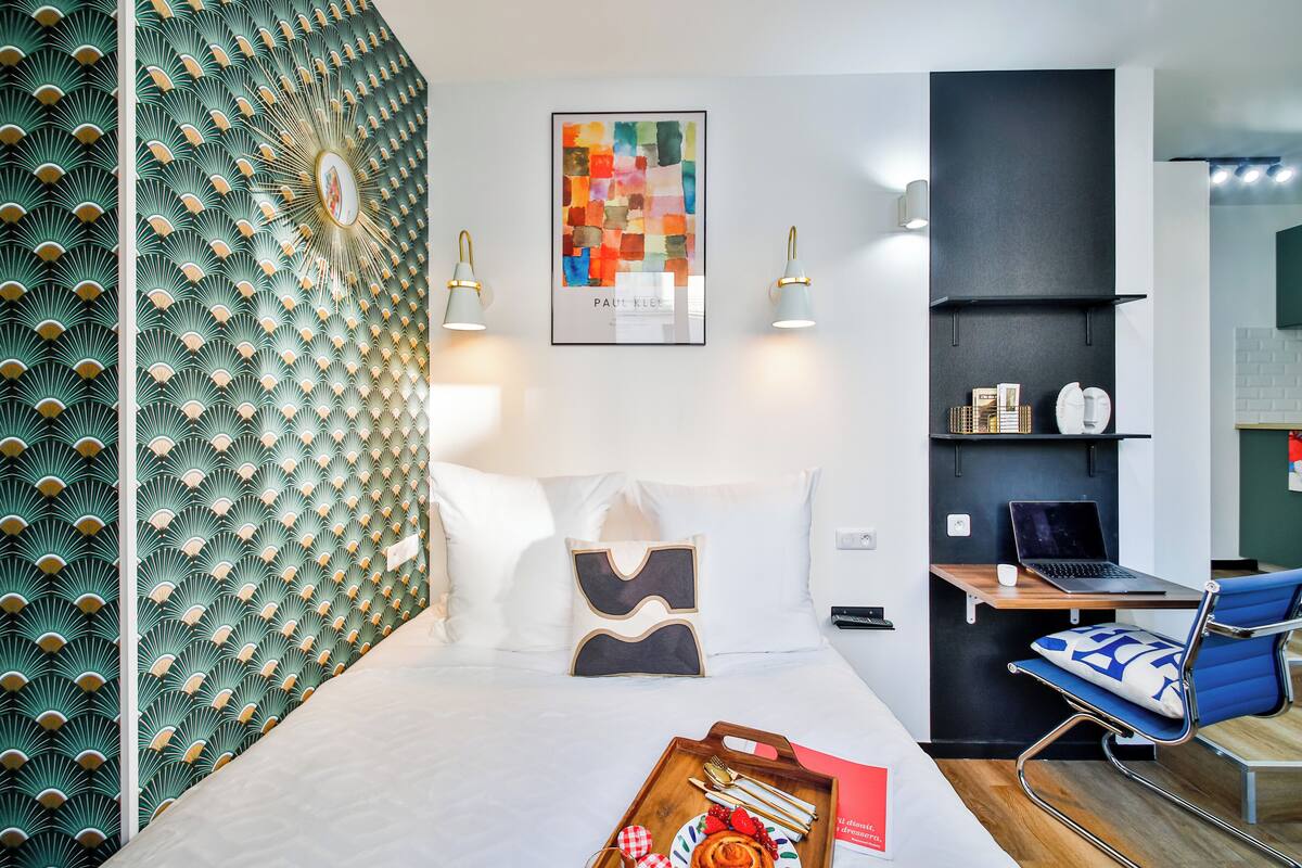Una habitación de hotel moderna con cama, escritorio con una computadora portátil, una silla azul y una bandeja con alimentos para el desayuno.