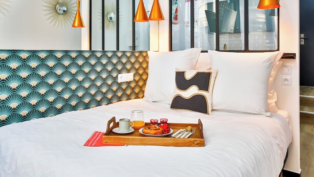 A neatly made bed with a tray of breakfast items, a modern headboard, and a large window with a view of the city. - WS Hôtel de Ville - Beaubourg (Paris)