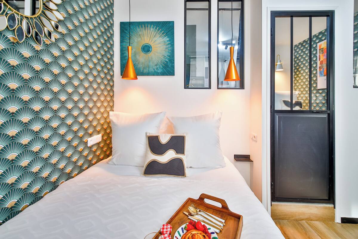 Una cama bien tendida con una bandeja de alimentos para el desayuno, una pared con un diseño geométrico y dos lámparas colgantes.
