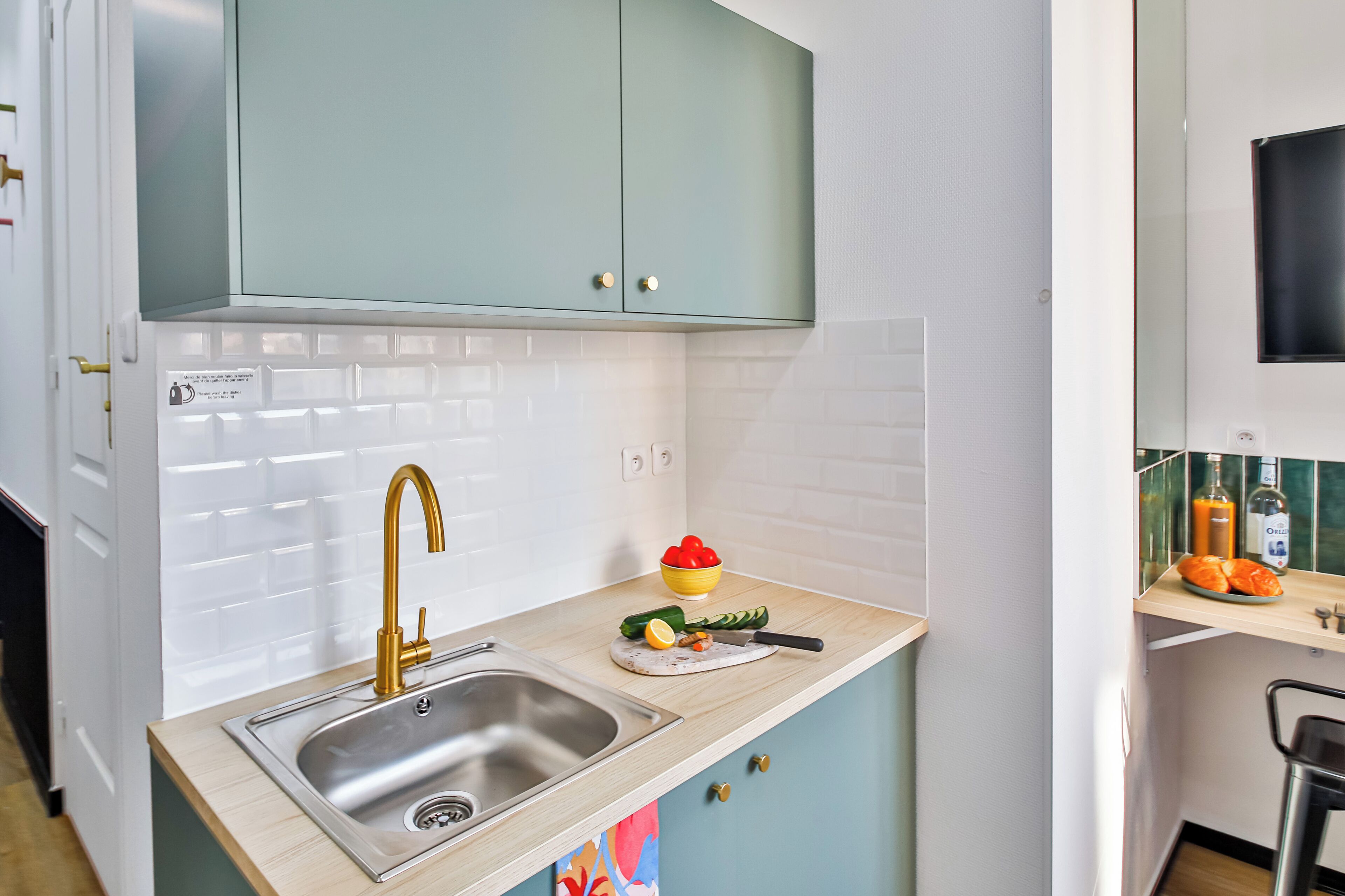 A kitchen with a stainless steel sink, a green cabinet above, and a countertop with a cutting board, knife, and vegetables.