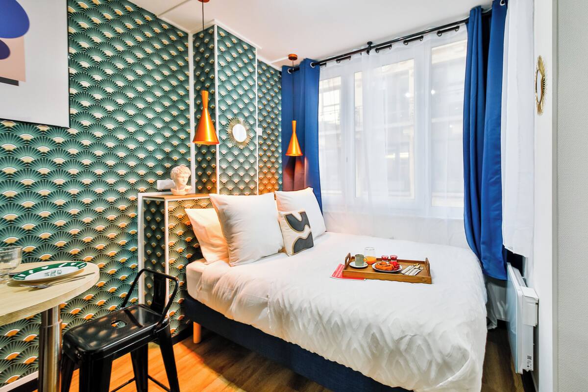 Una habitación de hotel ordenada con cama, una mesita con silla, un detalle decorativo en la pared y una ventana con cortinas.