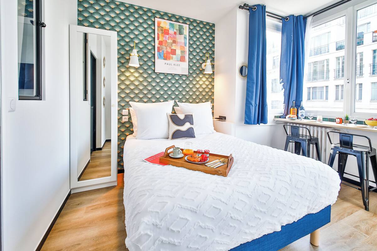 Una cama bien tendida con una bandeja de desayuno, un escritorio con sillas y vistas a la ciudad a través de amplios ventanales.