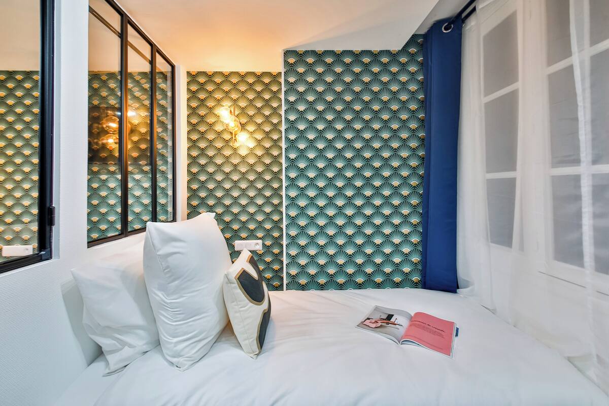 Una habitación de hotel con cama, almohadas, una revista y una ventana con cortinas.