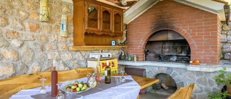 Un espace de restauration extérieur rustique, doté d’un four en briques, de murs en pierre, de meubles en bois et d’une table dressée avec de la nourriture et des boissons.