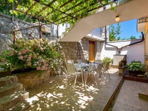 A stone-walled patio with a table and chairs, surrounded by potted plants and a pergola with vines.