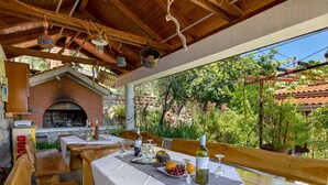Un espace de restauration en plein air avec des bancs et des tables en bois, un four en briques et une vue sur la verdure et un bâtiment doté d’un toit en tuiles.