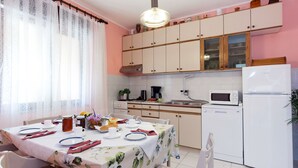 Un comedor con una mesa dispuesta para dos, una cocina con armarios y electrodomésticos blancos, y un ventanal con cortinas traslúcidas.