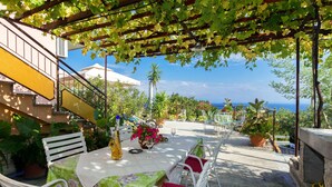 Un patio exterior con una mesa preparada para dos, rodeado de sillas blancas con cojines rojos, bajo una pérgola cubierta de enredaderas verdes.