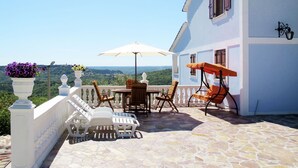 Eine Terrasse mit weißen Liegestühlen, einem Tisch und einem Sonnenschirm mit Blick auf eine schöne Landschaft.