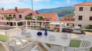 A balcony with a table set for two, white plastic chairs, a striped awning, and a view of buildings and mountains in the background. - Apartment Branka by Interhome (Rabac)