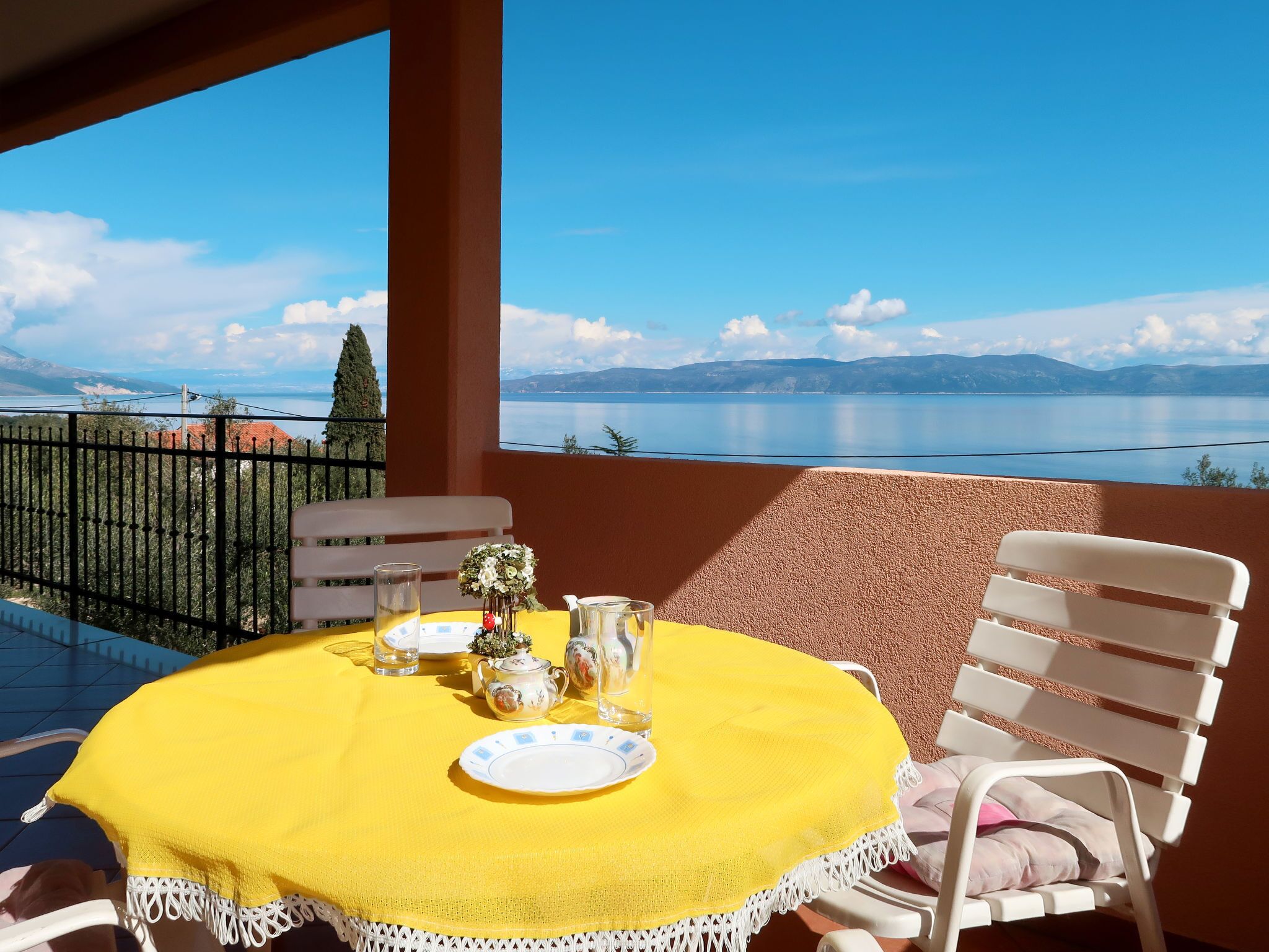 A balcony with a yellow table set for two, overlooking a lake and mountains.