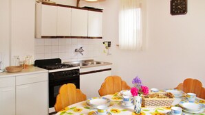 Cocina con armarios blancos, estufa y lavamanos. Se observa una mesa de comedor con mantel floreado y platos.