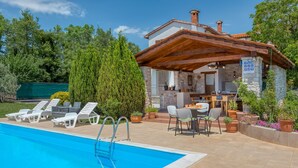 Zona junto a la piscina con sillones de descanso, una piscina y un edificio de piedra con una pérgola de madera.