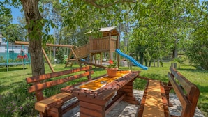 Una mesa de picnic de madera con bancos, un parque infantil con toboganes y columpios, y un árbol grande al fondo.