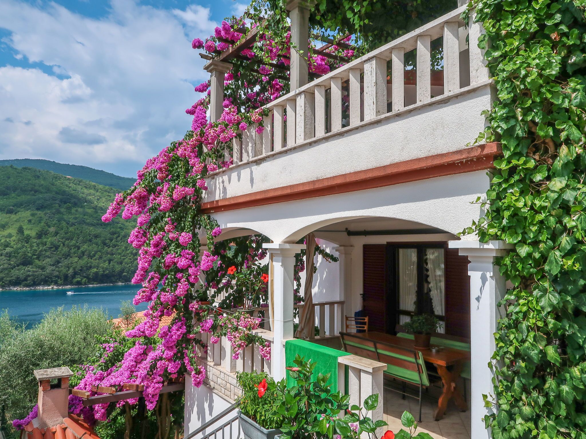 Ein zweistöckiges Gebäude mit einem Balkon, der in violette Blumen getaucht ist, mit Blick auf ein Gewässer und grüne Hügel.