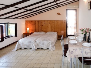 A bedroom with two beds, a wooden headboard, a nightstand with a lamp, a dining table set for two, and a window with curtains. - Violetta by Interhome (Labin)