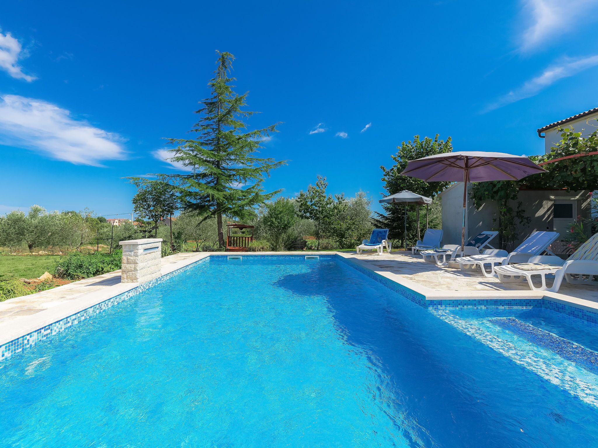 Piscina azul límpida com espreguiçadeiras e guarda-sóis, rodeada por vegetação e céu claro.