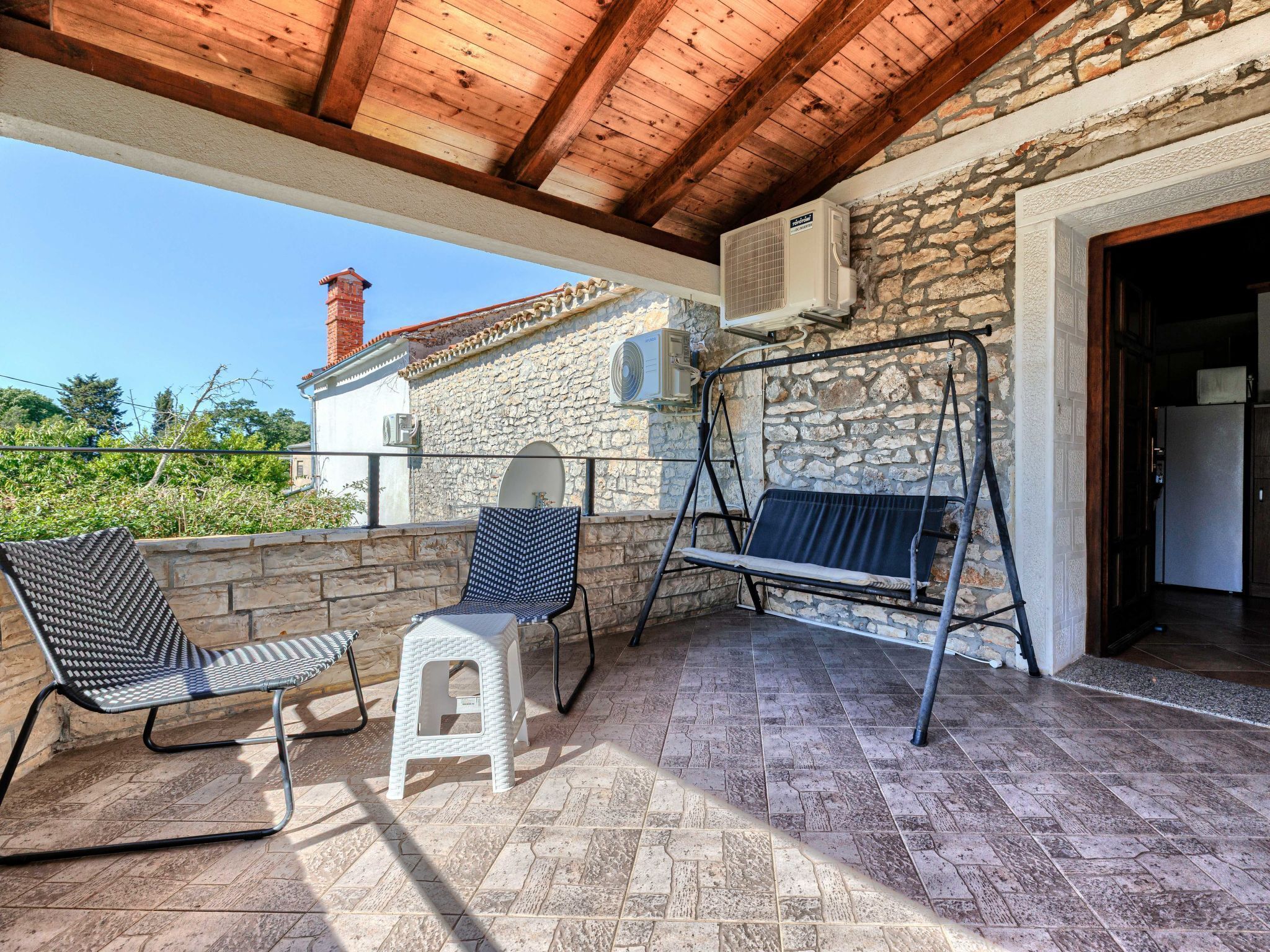 A covered patio with stone walls, a swing, two chairs, and an air conditioning unit.