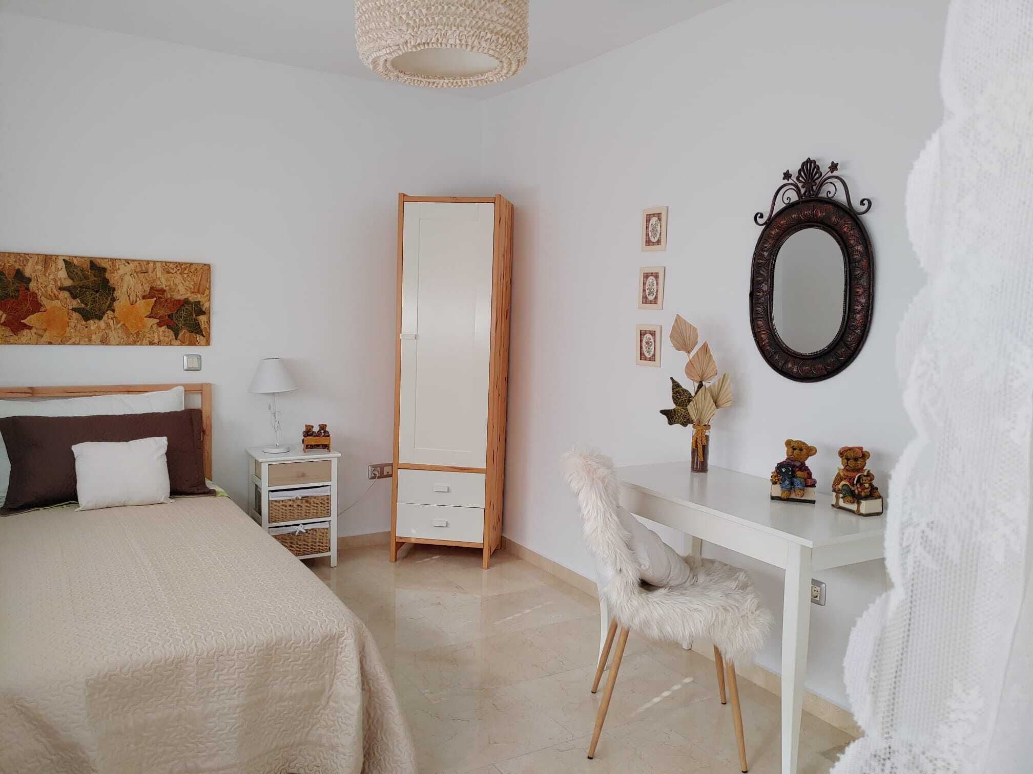 Un dormitorio con cama, cómoda, silla, escritorio, espejo y un muro con cuadros enmarcados.