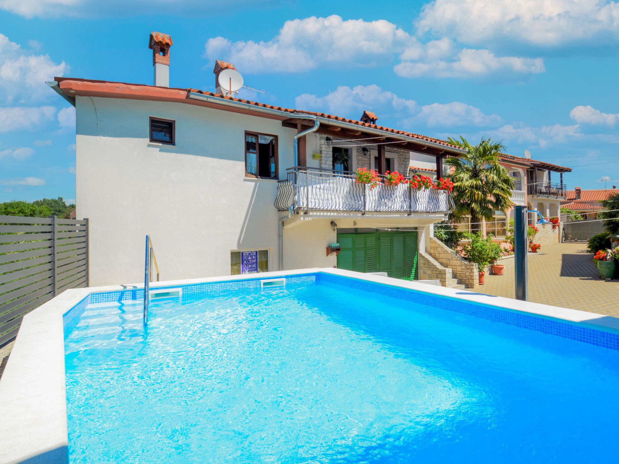 Vista da piscina de uma casa de dois andares, com uma piscina, uma varanda com flores vermelhas e um céu azul claro.