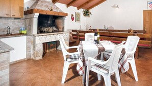A rustic kitchen with a stone fireplace, wooden beams, and a dining area with white plastic chairs and a table set for two. - Apartment Jukić by Interhome (Poreč/Kaštelir)