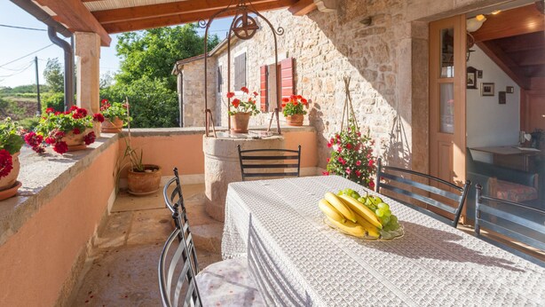 Un edificio de piedra con balcón, una mesa cubierta con mantel, sillas y macetas con flores.