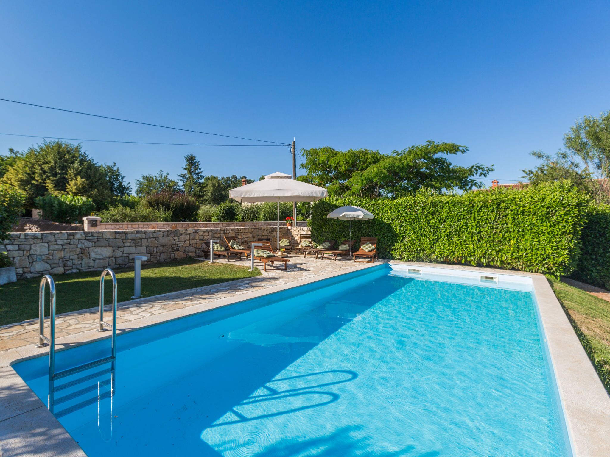 Una piscina rectangular con un muro de piedra y una escalera de piedra para entrar. Se ven sillones de descanso y sombrillas al fondo.
