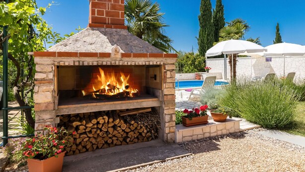 A brick outdoor oven with a fire burning inside, surrounded by a garden with plants and a pool in the background. - Apartment Koštro by Interhome (Poreč)