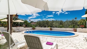 A swimming pool with lounge chairs and umbrellas, surrounded by greenery and a clear sky.