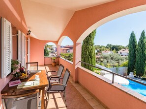 En balkon med et bord og stole, en potteplante samt udsigt over en swimmingpool og omkringliggende bygninger.