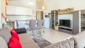 En moderne stue med sofa, stole, fjernsyn og et køkken med skabe og et køleskab.