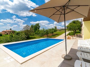 A swimming pool with a clear blue water, surrounded by a tiled deck, and an umbrella providing shade.