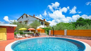 A swimming pool with lounge chairs and umbrellas, surrounded by a wooden fence and a house with a red roof. - Apartment Pina by Interhome (Pazin)