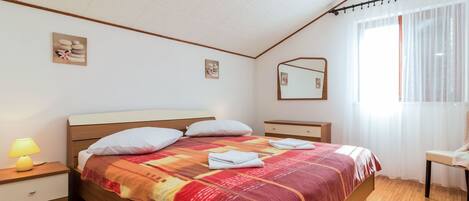 A bedroom with a wooden bed, a bedside table with a lamp, a small dresser, a mirror, and a window with sheer curtains.