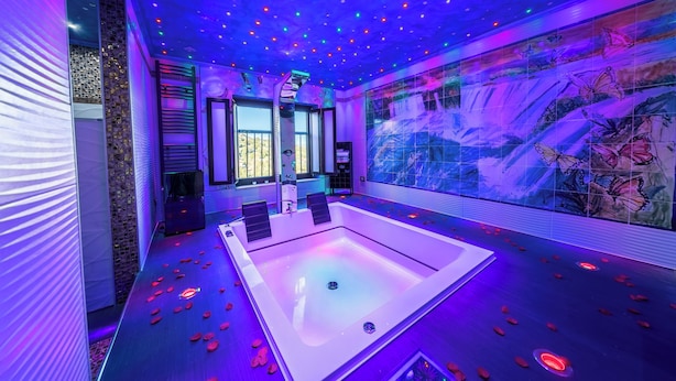 Moderni spa-huone, jossa on suuri, huoneen keskellä sijaitseva kylpyamme, värikkäillä valoilla varustettu katto ja seinällä suuri seinämaalaus, jossa on kuvattuna perhosia.
