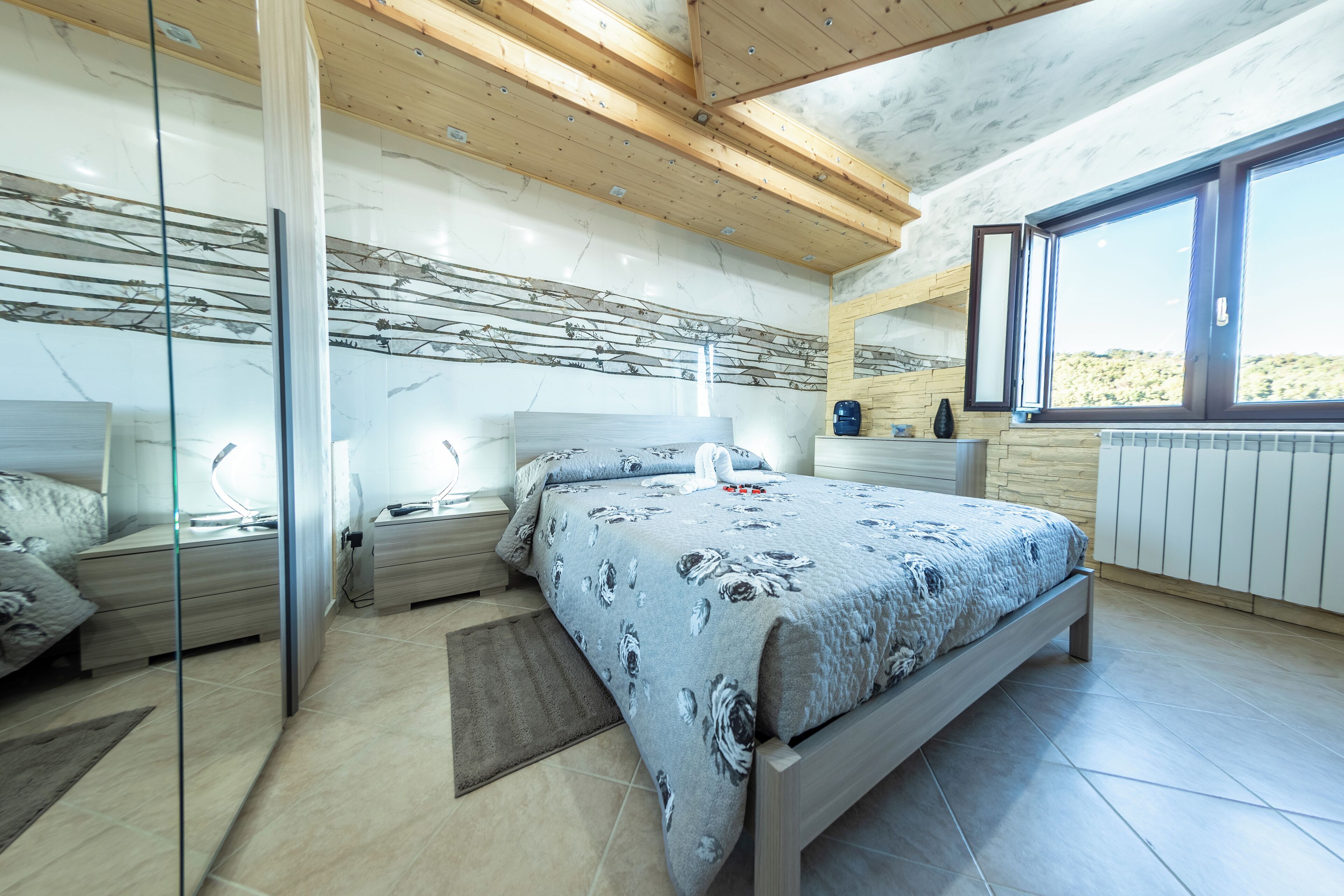 Un dormitorio moderno con una cama grande, mesitas de noche, un separador de vidrio, vistas al exterior y techo de madera.