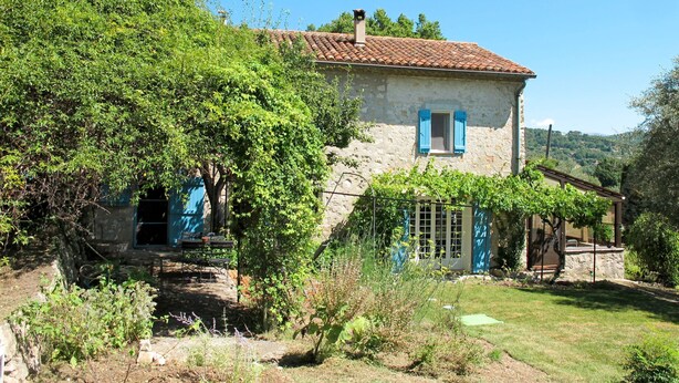 Une maison de campagne en pierre, avec un toit en tuiles, un petit porche et un jardin où poussent différentes plantes, ainsi qu’un banc.