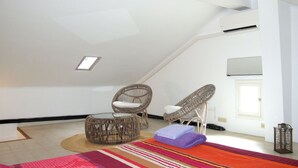 Une chambre spacieuse aménagée en mezzanine, avec un plafond en pente, deux fauteuils en osier, un petit pouf rond, un tapis coloré à rayures et une fenêtre laissant entrer la lumière naturelle.