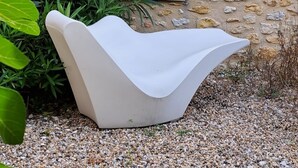 Ein weißer, moderner Stuhl in einem Garten mit einer Steinmauer und einem Fenster mit Fensterläden.