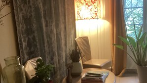 Ein gemütliches Wohnzimmer mit Couchtisch aus Holz, einem weißen Sessel, einem kleinen Beistelltisch mit Vase und Pflanze sowie einem Fenster mit Vorhängen.