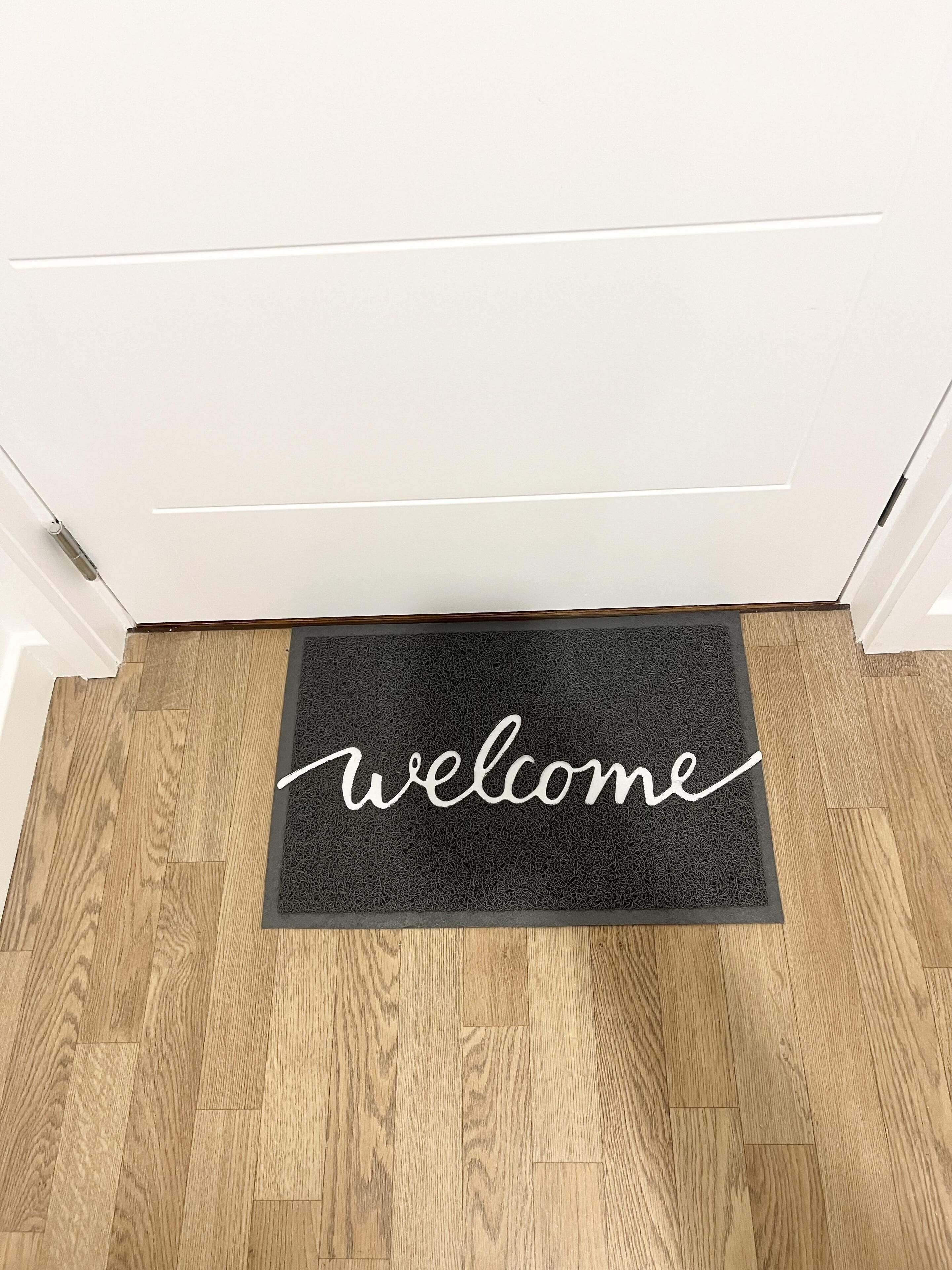Un tappetino nero con la scritta 'welcome' in corsivo su un pavimento di legno.