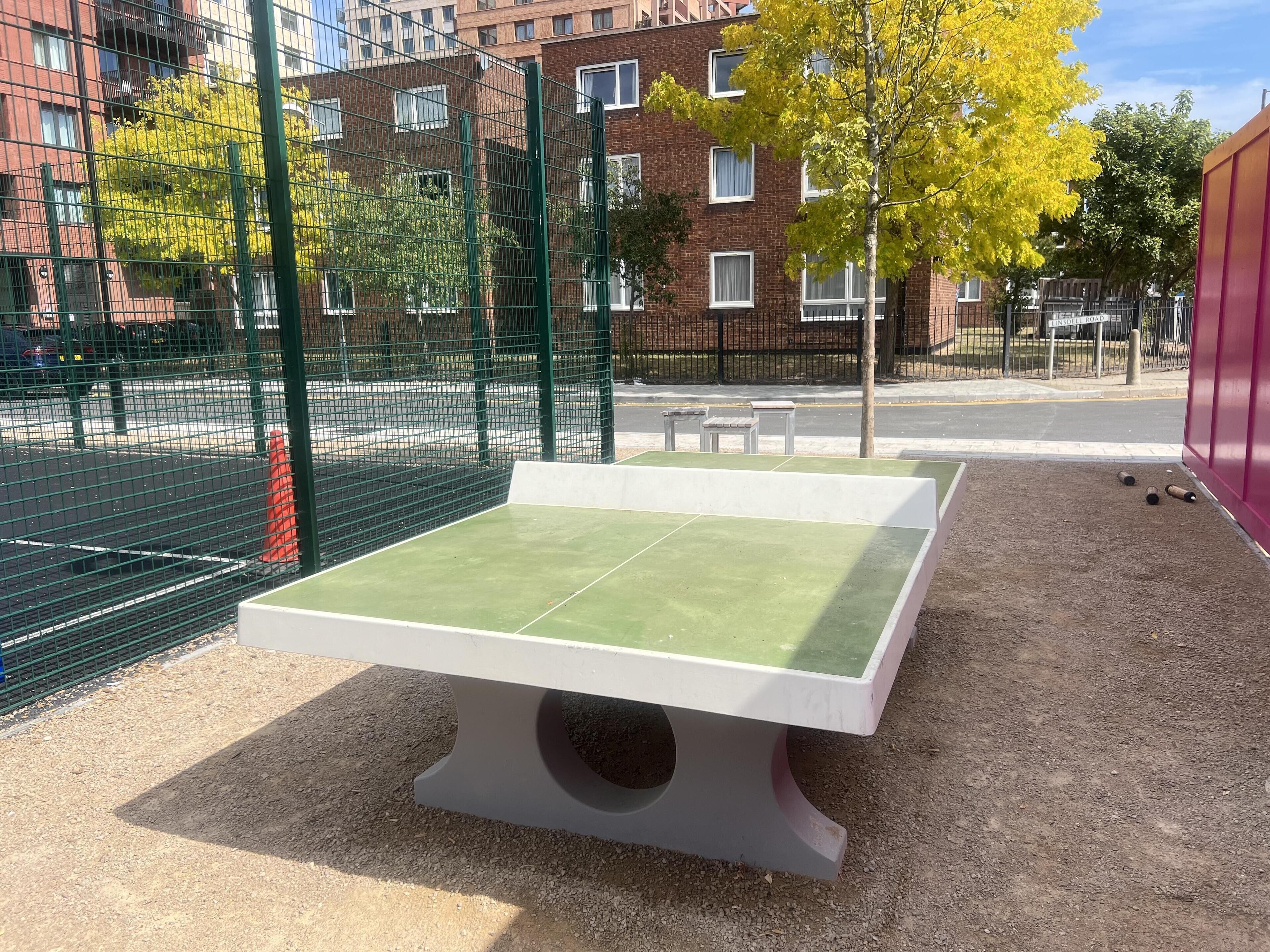 A ping pong table in an outdoor area with a fence, gravel ground, and buildings in the background.