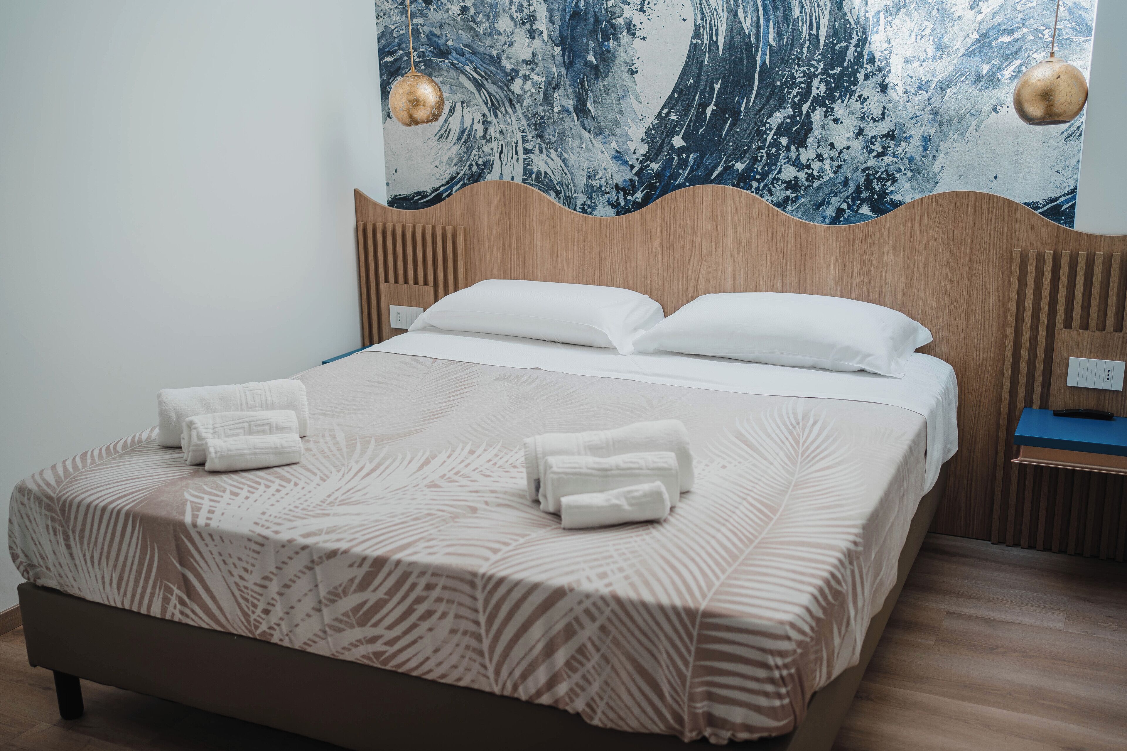 A neatly made bed with a leaf-patterned bedspread and two rolled towels, a wooden headboard, and a wall with a large abstract painting.