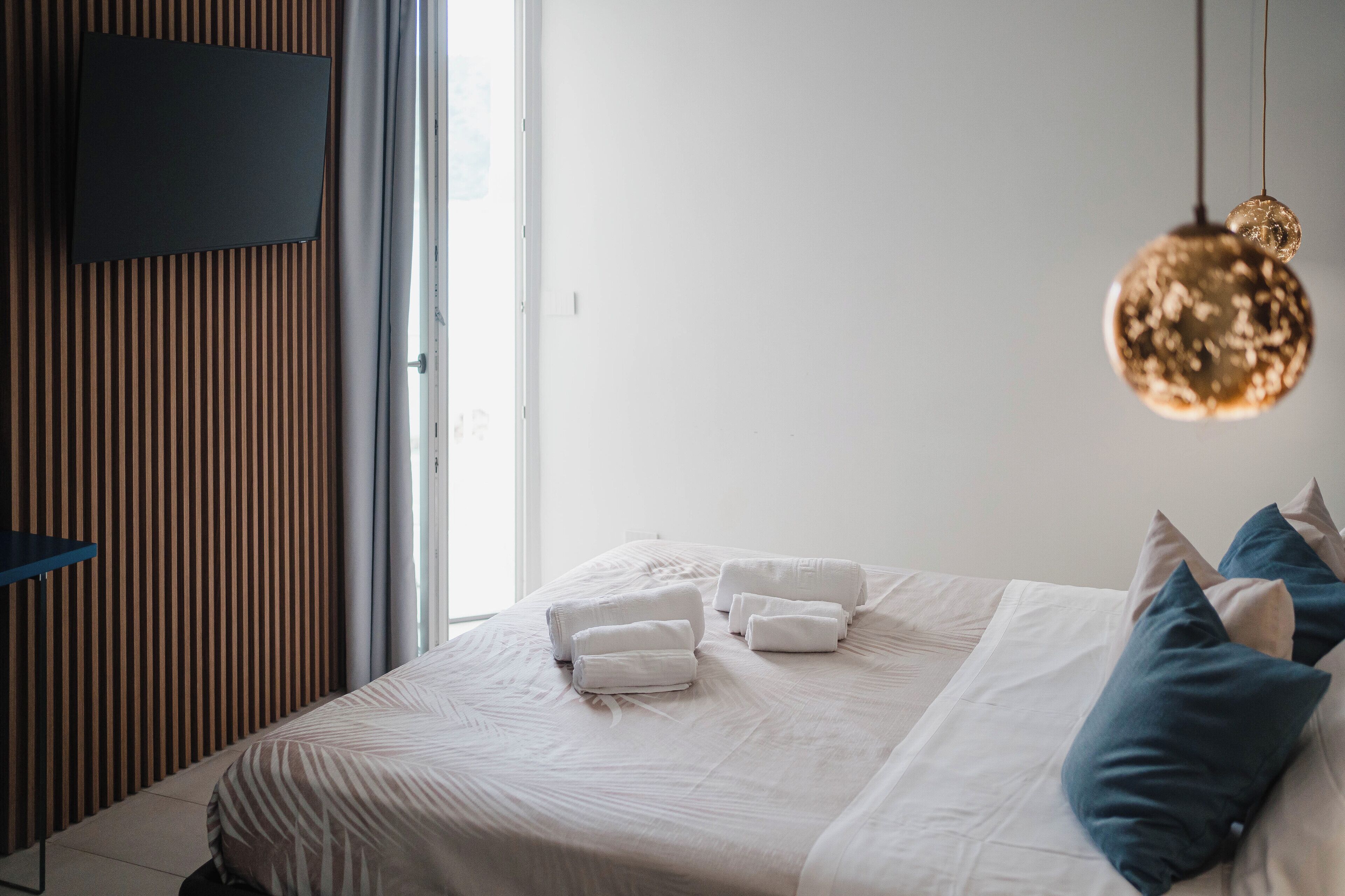 A neatly made bed with white linens and rolled towels, a television mounted on a wooden panel wall, and a hanging light fixture.