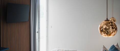 Una cama bien tendida con sábanas blancas y toallas enrolladas, un televisor montado en una pared de paneles de madera, y una lámpara colgante.