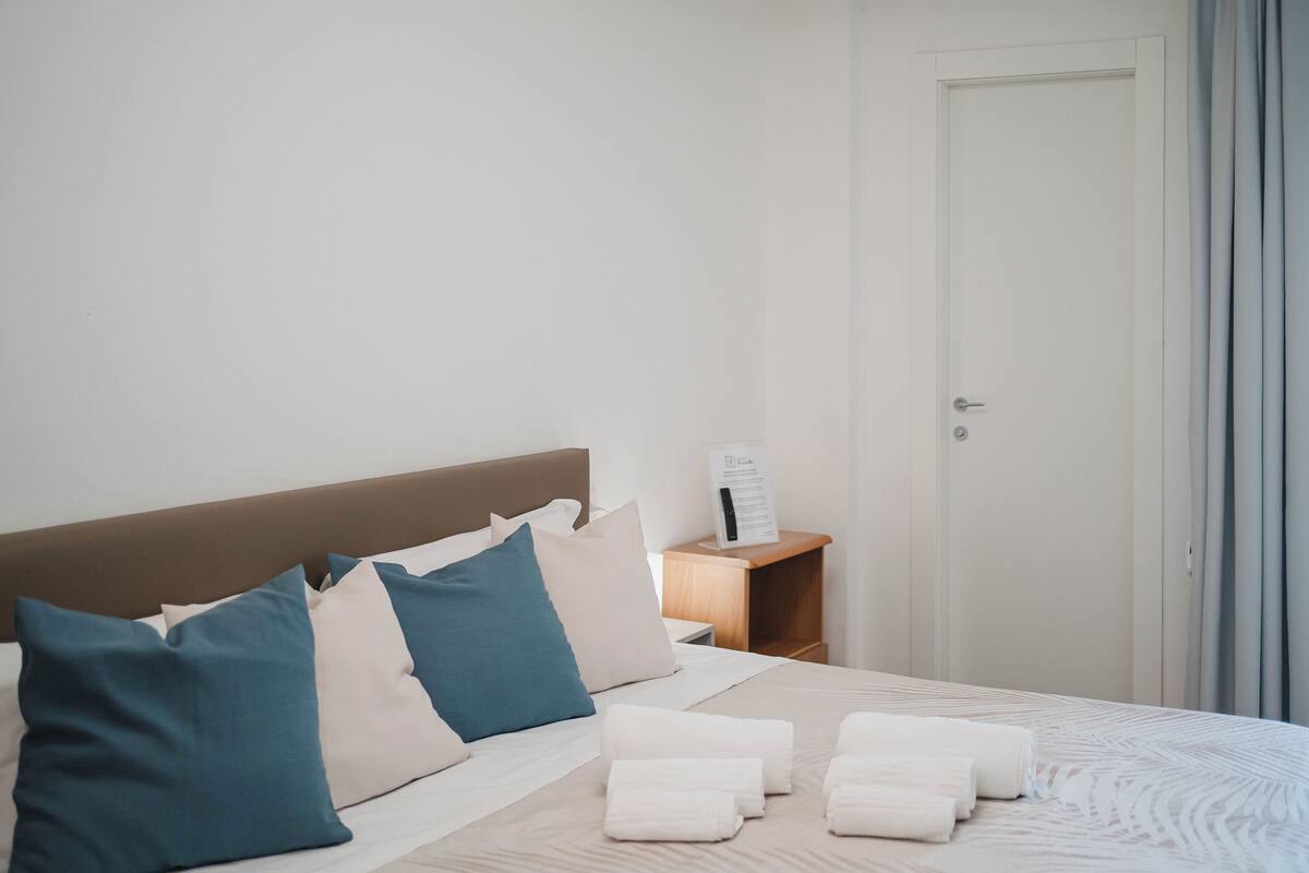 Ein ordentlich bezogenes Bett mit Kissen, ein Nachttisch mit Lampe und eine Tür mit Vorhängen.
