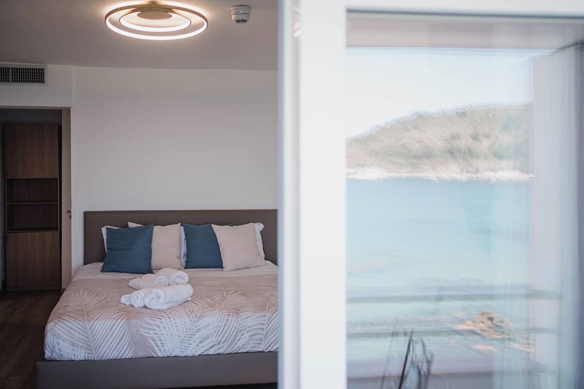 Ein modernes Schlafzimmer mit Bett, Meerblick durch ein großes Fenster und einer Deckenleuchte.